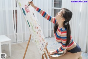 A woman sitting on a stool painting on a easel.